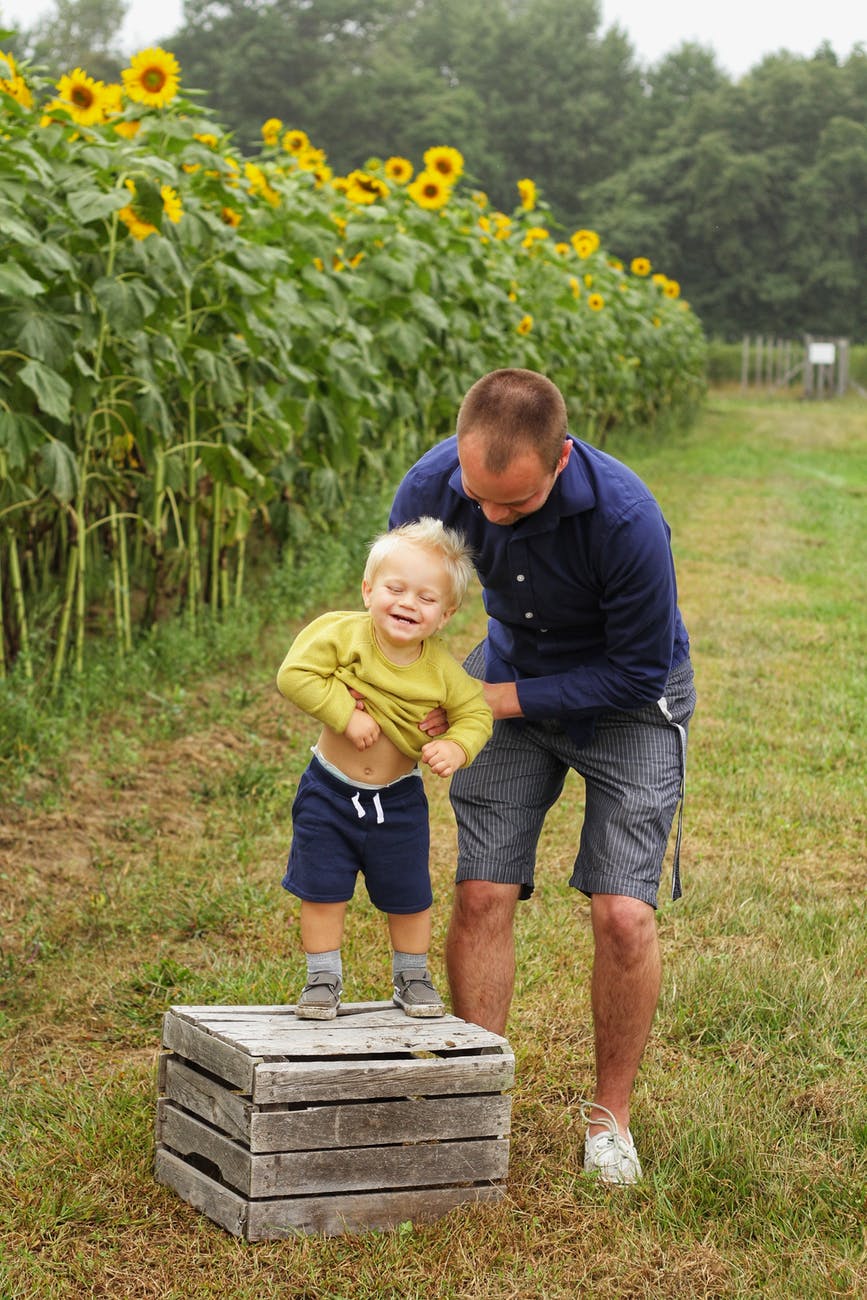 man holding smiling child standing on brown wooden crate near sunflowers